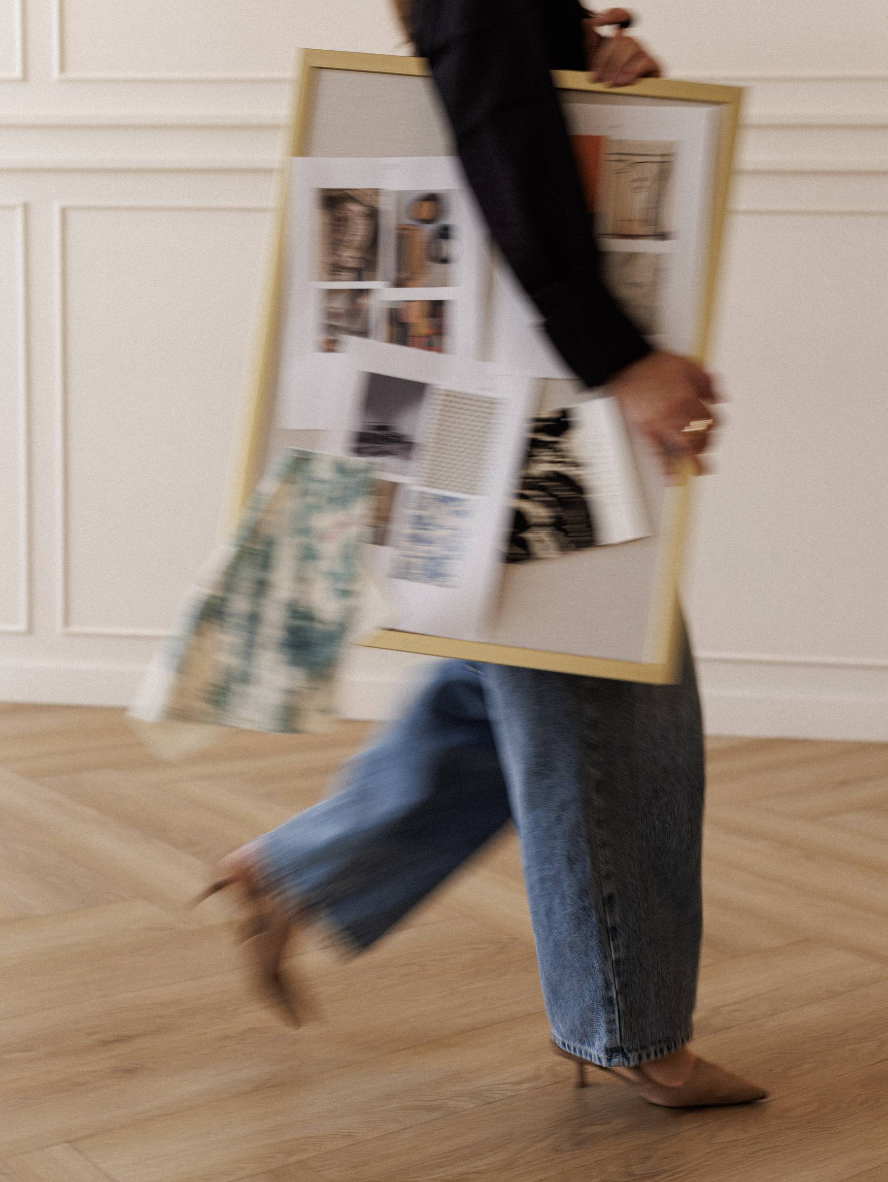 Carrying a framed mood board of reference images across a room with panelled walls and a herringbone floor